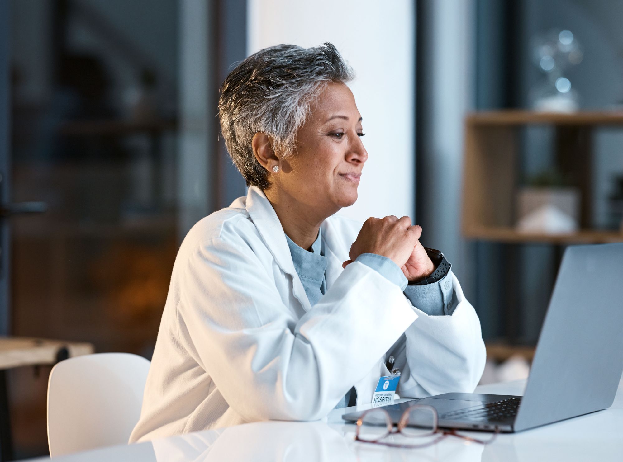 Healthcare professional in a white coat sits at a laptop, hands clasped, in a clinical setting.
