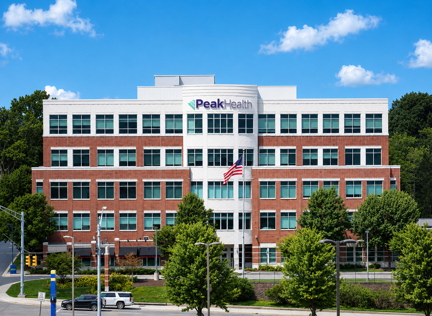 Front view of the Peak Health office building, a red-brick with white facade, large windows, and an American flag in front under a blue sky.