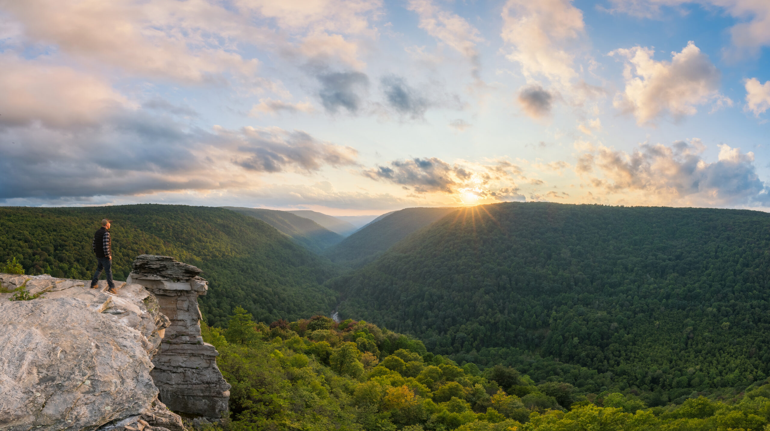 Person stands on a rocky cliff edge overlooking a dense forested valley at sunset.